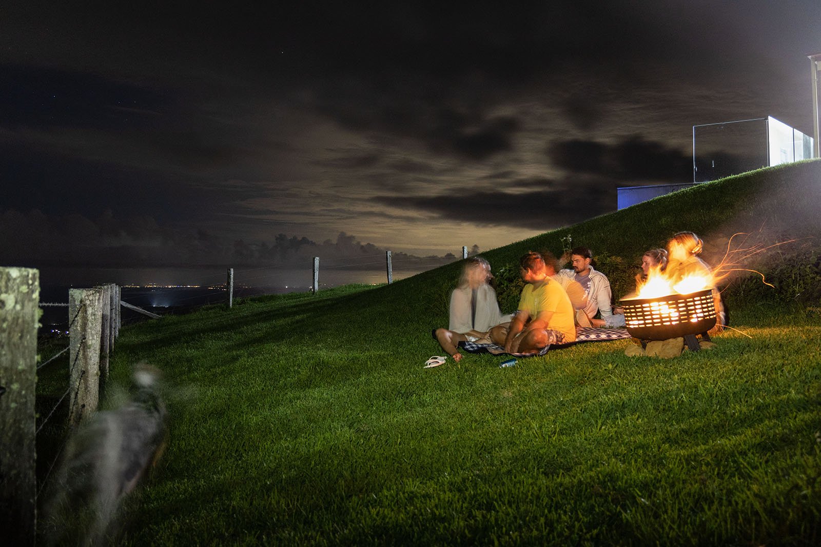 Gather around a glowing fire pit on a grassy hill under a starry night sky, with a dimly lit horizon in the background.
