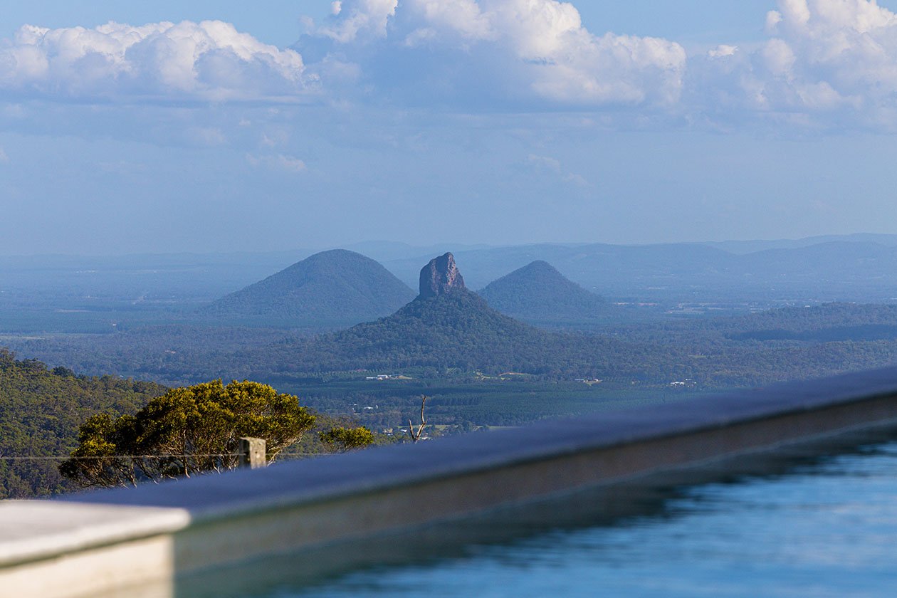 View of the Glass House Mountains' three peaks in the distance with a swimming pool edge in the foreground, and the lush rainforest surrounding.