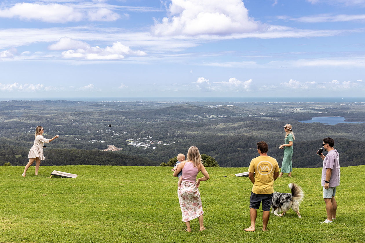 Play outside on the Sunshine Coast hinterland grassy hill with a scenic view of towns, forests, and coastlines below under a cloudy blue sky.